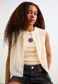 Cardigan senza maniche in maglia crema sopra un top in cotone beige a costine, abbinato a pantaloncini in denim neri a vita alta. Presenta una toppa rotonda sul top.