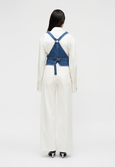 Γυναίκα με μακριά σκούρα μαλλιά που φοράει λευκό παντελόνι φαρδιάς γραμμής, λευκό πουκάμισο με μακριά μανίκια και μπλε κοντό τζιν γιλέκο με ιμάντες στην πλάτη.