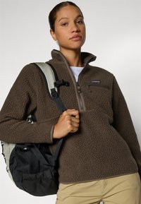 Sudadera de forro polar marrón con cuello alto, bolsillo en el pecho con cremallera y superficie texturizada. Combinada con una mochila ligera y pantalones beige.