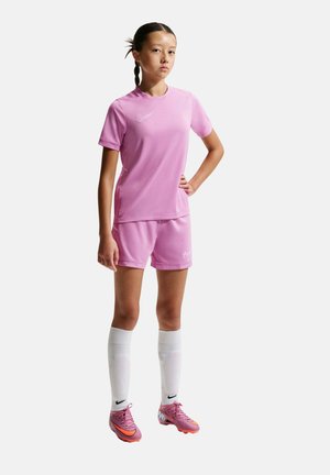 Joven atleta de pie con la mano en la cadera, vistiendo ropa deportiva rosa, calcetines blancos hasta la rodilla y tacos morados sobre un fondo liso.
