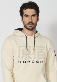Sudadera beige hecha de una tela suave con un bolsillo frontal. Presenta cordones oscuros, un logotipo texturizado y letras negras de "KOROSHI".