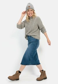 Grå strikket sweater med kabelmønster, blå denim midi-nederdel og brune ankelstøvler. Model iført grå strikket beanie, smilende.
