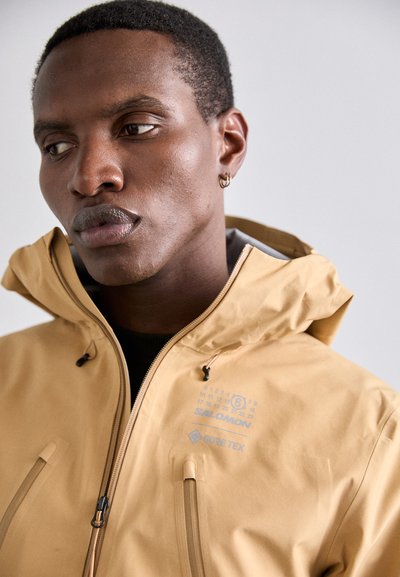 Veste Gore-Tex jaune avec fermeture éclair, capuche réglable et deux poches avant. Comprend un logo imprimé et des éléments de design numériques.