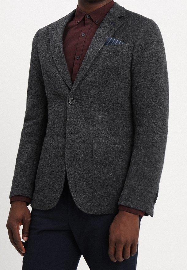 Uomo che indossa un blazer grigio scuro a trama sopra una camicia bordeaux con bottoni, un fazzoletto da taschino blu navy e pantaloni scuri.