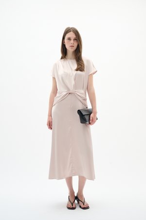 Kobieta w jasnoróżowej sukience midi z ozdobnym wiązaniem w talii, trzymająca czarną kopertówkę i nosząca czarne sandały na obcasie na białym tle.