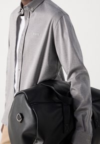 Grijze button-up shirt met een textuurpatroon en geborduurd logo, gecombineerd met een grote zwarte leren sporttas met afgeronde hoeken en een metalen accent.