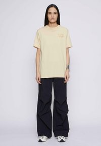 Camiseta de algodón crema con mangas cortas y escote redondo, que presenta el texto "DO NOTHING Club" bordado en naranja. Lleva pantalones negros holgados.