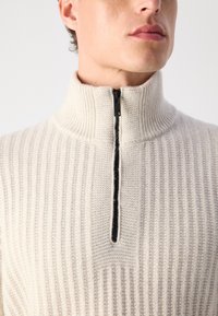 Cremfarbener Strickpullover mit hohem Kragen, halber Reißverschluss und vertikaler Rippstruktur mit einem kontrastierenden Reißverschlussdetail.