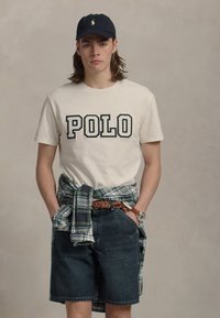 Jauns vīrietis, ģērbies melnā cepurē, krēmkārtā POLO krekls, plānas svītru krekliņš sasietas ap jostasvietu, džinsu šorti un rokas pulkstenis, stāv ar rokām kabatās.