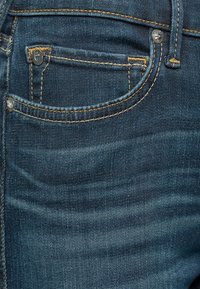 Granatowe dżinsy w klasycznym pięciokieszeniowym wzorze z kontrastującymi żółtymi przeszyciami oraz widocznym guzikiem i rozporkiem z przodu.