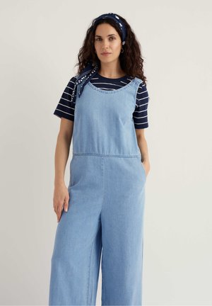 Nainen, jolla on kiharat tummat hiukset, pukeutunut laivastonsiniset raidalliseen paitaan, vaaleansinisiin farkkuhaalarit ja laivastonsininen pilkullinen hiuspanta, yksinkertaisen taustan edessä.