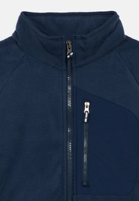 Tamsiai mėlynas vilnonis džemperis su aukštu apykakliu, priekiniais užtrauktuku ir užtrauktuku užsegama krūtinės kišenė. Charakterizuojamas lygiu audiniu ir kontrastine audinio plokštele.