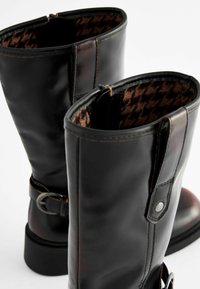 Schwarze Lederstiefel bis zum Knie zeichnen sich durch eine glatte Oberfläche, einen verstellbaren Schnallenriemen und ein Futter im Glencheck-Muster aus.
