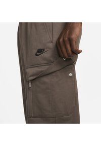 Ruka upravující patentku na hnědých kalhotách Nike s kapsou, viditelným zipem a černým logem Nike.