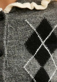 Grå stickad cardigan med ett svart och vitt argyle-mönster. En rynkad krämfärgad krage med svarta knappar ger extra detaljer åt designen.