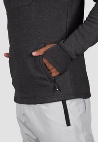 Pullover grigio scuro con tasca laterale, polsini a costine e una texture liscia, abbinato a pantaloni grigi chiari con tasche con zip.