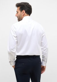 Weißes Langarmshirt mit klassischem Kragen, schlanker Schnitt und knopfbaren Manschetten, kombiniert mit dunklen marineblauen Hosen mit Gesäßtaschen.
