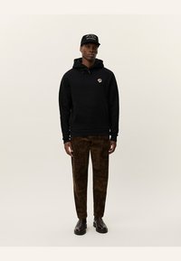 Svart hettegenser med snor og logo, kombinert med brune mønstrede bukser. Modellen har på seg svart caps og brune ankelstøvler.