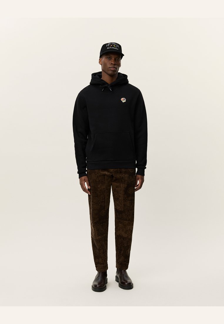 Svart hettegenser med snor og logo, kombinert med brune mønstrede bukser. Modellen har på seg svart caps og brune ankelstøvler.