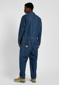 Combinaison en denim bleu foncé, avec un col, des manches longues et des poches arrières. Détails de couture notables et étiquette de marque visibles.