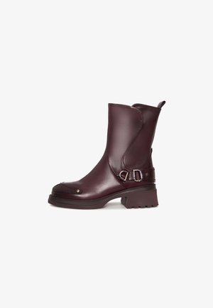 Stivaletto in pelle marrone scuro con tacco basso e spesso, fibbia decorativa sul lato e linguetta sul retro, mostrato di profilo.