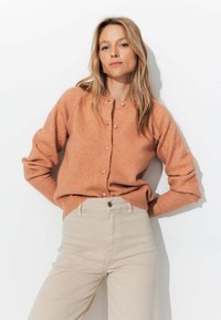 Cardigan en tricot de couleur pêche avec un col rond, fermeture à boutons et poignets côtelés, associé à un jean taille haute beige clair.