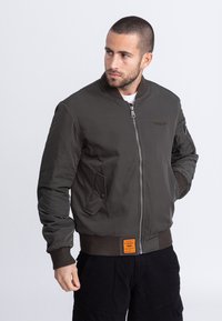 Kurtka bomberska w ciemnooliwkowym kolorze, nylonowa tkanina, ściągacz na kołnierzu i mankietach, zamek błyskawiczny z przodu, boczne kieszenie, pomarańczowe detale z etykietą.