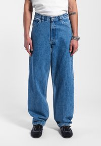 Jeans de mezclilla azul holgados con una cintura alta, presentan bolsillos grandes y una textura sutil. Combinados con zapatos negros y una camisa blanca.