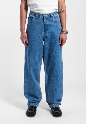 Baggy blauwe spijkerbroeken met een hoge taille, met grote zakken en een subtiele textuur. Gestyled met zwarte schoenen en een wit shirt.