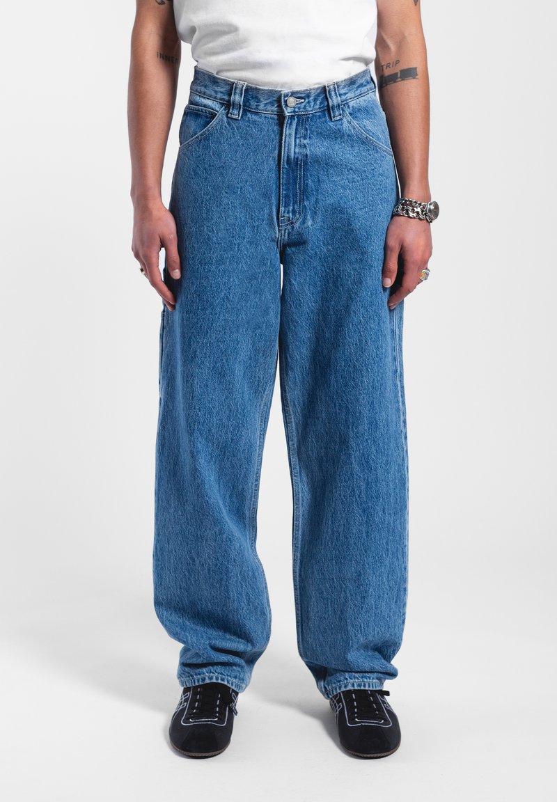 Baggy blå denimjeans med hög midja, stora fickor och en subtil textur. Stiliserade med svarta skor och en vit skjorta.