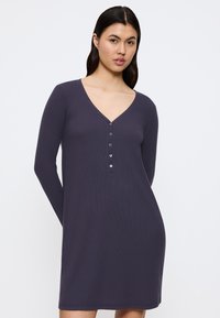 Donna con lunghi capelli scuri che indossa un vestito a maniche lunghe, viola scuro, con scollatura a V e allacciai a bottoni, in piedi davanti a uno sfondo bianco semplice.