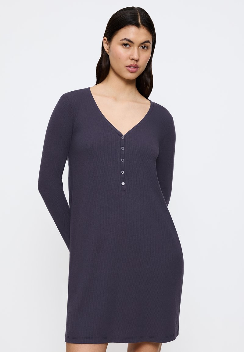 Donna con lunghi capelli scuri che indossa un vestito a maniche lunghe, viola scuro, con scollatura a V e allacciai a bottoni, in piedi davanti a uno sfondo bianco semplice.