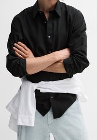Camicia nera con bottoni e maniche lunghe, dal taglio morbido, con una camicia bianca annodata in vita, indossata con pantaloni azzurri chiari.
