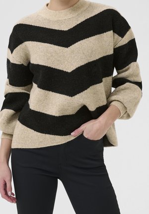 Persona vistiendo un suéter beige y negro con rayas en forma de chevron y mangas abullonadas, combinado con pantalones negros, con la mano en el bolsillo.