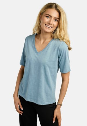 Plavolaska ženska se smehlja, nosi svetlo modro majico z V-izrezom s kratkimi rokavi in črne hlače, stoji z roko na boku pred enobarvnim ozadjem.