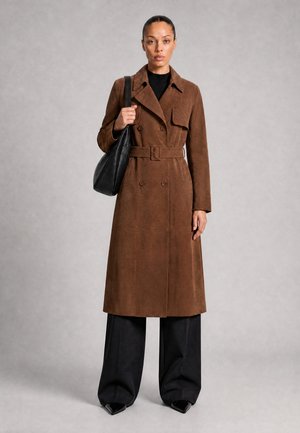 Kvinna som bär en brun trenchcoat med bälte, svarta vida byxor och håller en svart axelväska mot en enfärgad bakgrund.