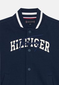 Námornícky modrá varsity bunda s bielym a námornícky pruhovaným golierom, vyšitým logom "HILFIGER" a predným zapínaním na gombíky. Mäkka, textúrovaná tkanina.