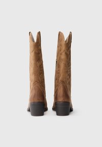 Ein Paar braune Wildleder-Cowboy-Stiefel mit dekorativer Stickerei und gestapelten Absätzen, von hinten auf weißem Hintergrund betrachtet.