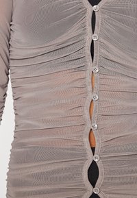Graues Mesh-Top mit geraffter Textur, ausgestattet mit einer vorderen Knopfverschluss, das eine hautfarbene Unterlage und dekorative Knopfakzente zeigt.