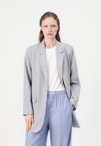 Grå blazer med klassisk slag och två framfickor, kombinerad med vit T-shirt och blå randsydda byxor. Slät tygkvalitet och skräddarsydd passform.