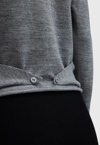 Szary sweter o fakturowanym wykończeniu z dwoma szarymi guzikami przy brzegu. Połączony z czarnymi prążkowanymi legginsami, podkreślającymi kontrast tkaniny i koloru.
