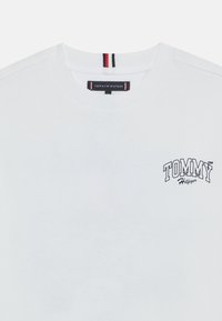 Hvit bomulls t-skjorte med rund hals, med en svart "TOMMY HILFIGER" logo på venstre bryst og stripete detaljer på kragen.