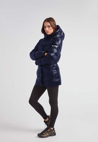 Mørkeblå puffjakke med hætte, skinnende materiale, indsnævret talje og lynlås-lommer. Båret over sorte leggings og sportssko med guld-detaljer.