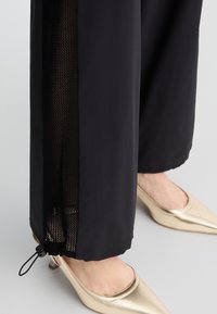 Pantaloni neri con pannelli laterali in rete, orlo elastico e dettaglio con cordoncino. Abbinati a scarpe slip-on lucide dorate.