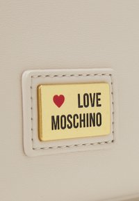 Beige läder med en guldfärgad rektangulär plack som har ett rött hjärta och svart text "LOVE MOSCHINO" inramat av sömnad.