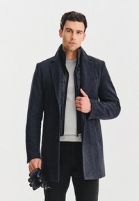 Czarny wełniany płaszcz z wyciętym kołnierzem, kieszonką z przodu i wnętrzem na suwak. W połączeniu z szarym swetrem i czarnymi rękawiczkami.