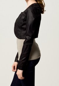 Svart kort jacka med knapplikning fram och två bröstfickor; bärs över en ljusgrå topp, i kombination med mörka jeans. Slät textur.