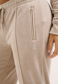 Beige sweatpants van soepel materiaal, met twee ritszakken en een decoratieve logo in strass op de bovenbeen.