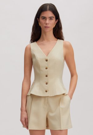 Frau trägt ärmellose beige Weste mit Knöpfen und passenden hoch taillierten Shorts, stehend vor einem schlichten hellen Hintergrund.