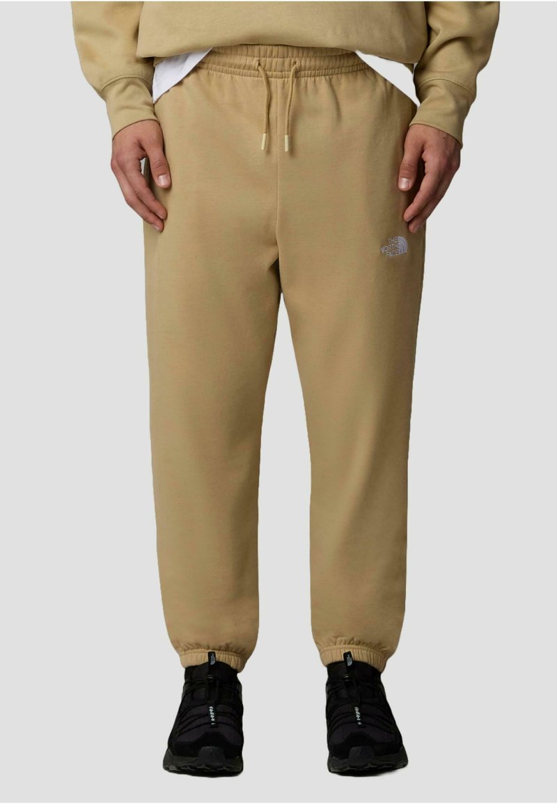 Beiga joggingsbyxor gjorda av tyg med en mjuk textur, med ett elastiskt midjeband och mudd i bensluten. En liten logotyp är synlig på vänstra låret.